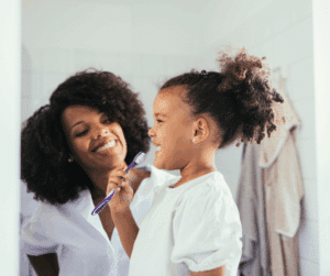 A happy young girl with curly hair holds a purple toothbrush, smiling with her mother in the mirror.