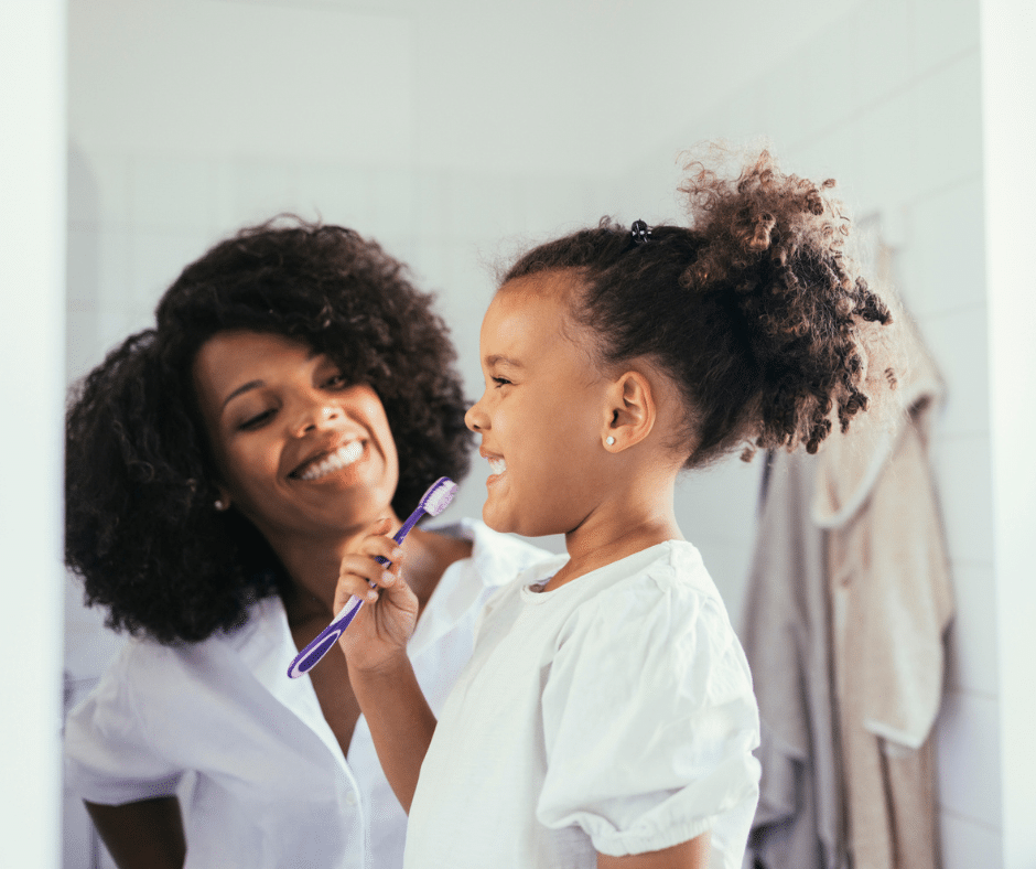 A happy young girl with curly hair holds a purple toothbrush, smiling with her mother in the mirror.