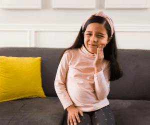A young girl in a pink shirt sits on a couch, wincing in pain and holding her hand to her cheek, indicating a toothache.